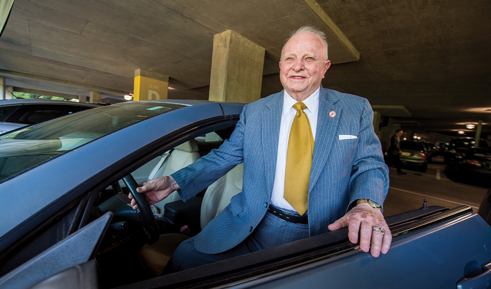 A man wearing a suit and tie stands next to a car with one hand on the steering wheel and one hand on the driver’s side door